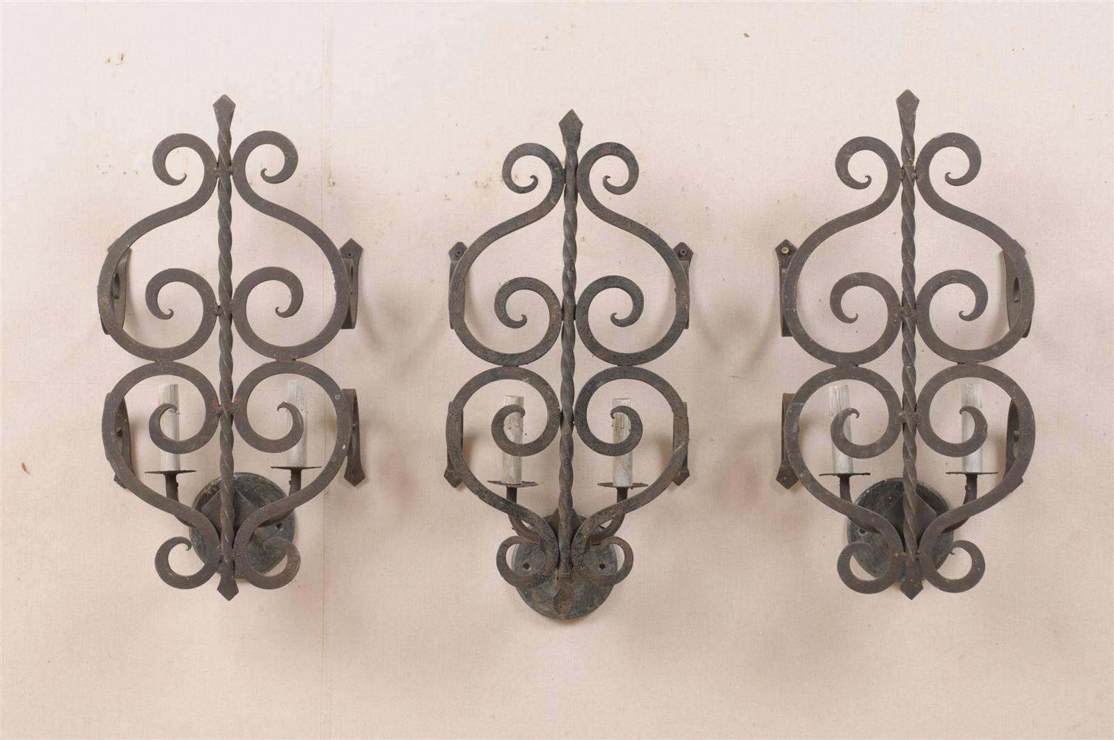 A set of three French two-light forged iron sconces with unusual projection of the iron work over the lighting area. The metal work features a theme of S-scrolls flowing symmetrically on each side of the central arm.  This set of three lovely French