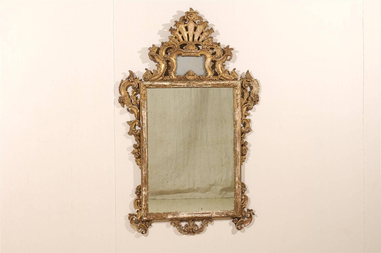 An Italian 19th century mirror. This giltwood Rococo style Italian mirror features a highly carved crest with flower carving at the very top. Below the carving, a cartouche motif. A series of volutes and scrolls decorate the sides. The corners of