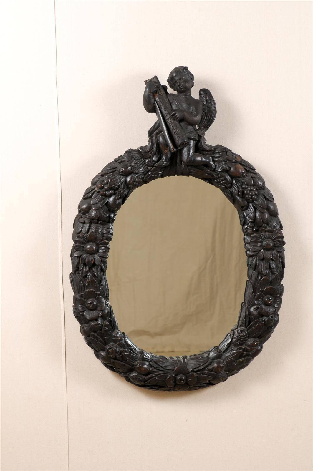 An 18th century English medium sized oval mirror. This English wall mirror has a carved wood crest featuring an angel holding a manuscript. The frame depicts a wreath of floral and fruit motifs.  This piece is a rich dark brown, almost black color