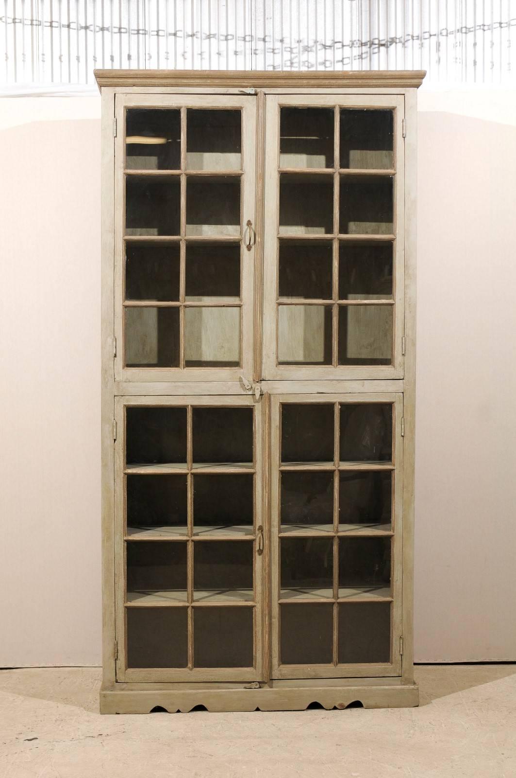 A tall, painted wood glass doors cabinet. This painted wood cabinet from India features a linear profile with four glass muntins doors. The doors open up to multiple inner shelves. The cabinet rests on a slightly scalloped base. The cabinet is of a