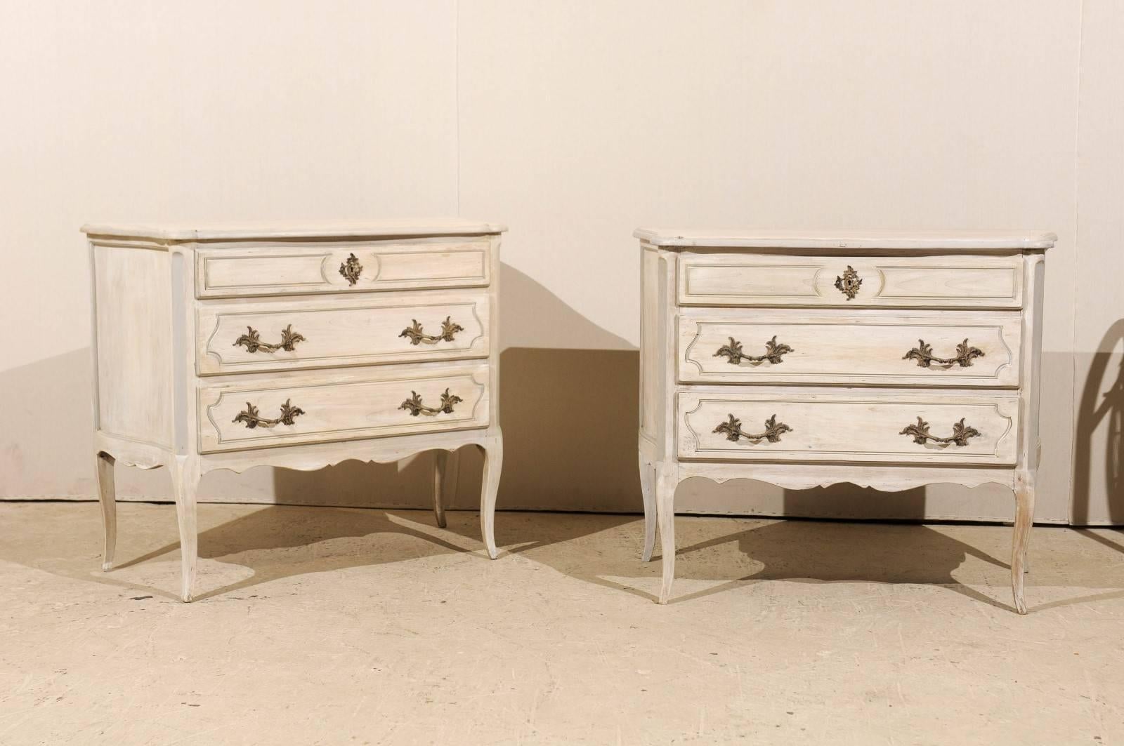 A pair of American vintage three-drawer chests. This pair of painted wood chests features a smaller drawer at the top with Rococo style central escutcheon over two larger drawers. The curvy top, cabriole legs and scalloped skirt on the three sides