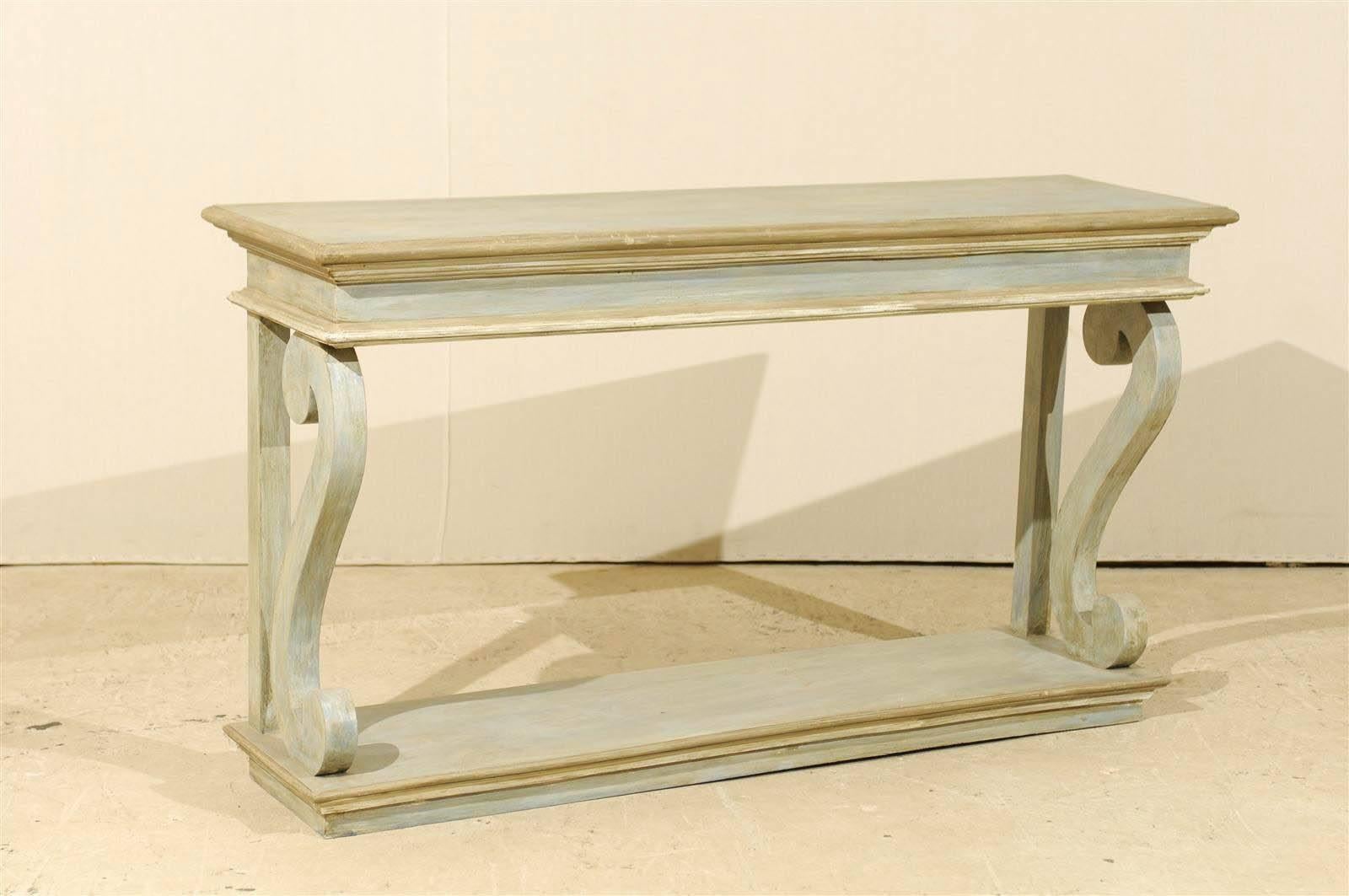 One hand carved painted wood Brazilian console made of reclaimed wood with voluted supports and lower tier. Nice clean look balanced by the elegance of the volutes. The color is a mix of blue, grey and green. The trim is more of a taupe hue. We have