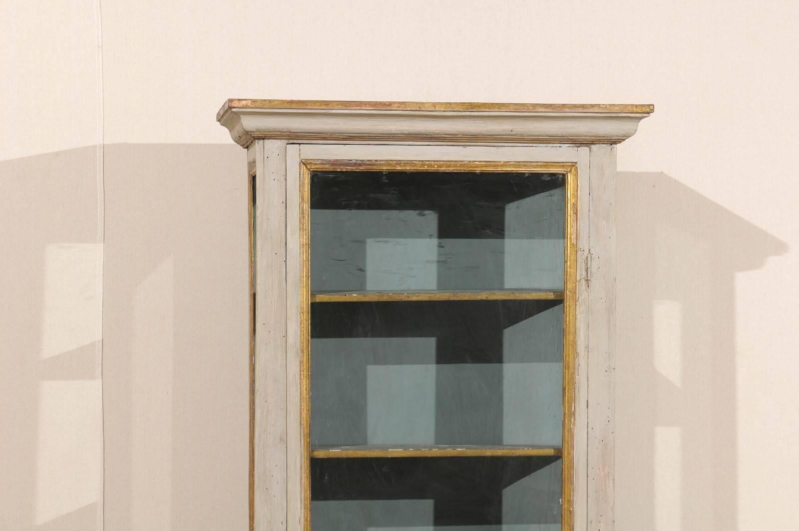 An Italian, early 19th century shallow display cabinet. This lovely one door cabinet features a linear profile with clean lines and is raised on tapered legs. It has its original grey color with gilded accents on the trim and blue painted interior