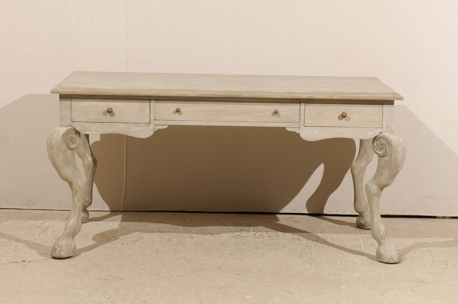 A painted wood desk with unique carved animal legs. This wooden desk features three drawers over four animal legs with hooved feet and scroll design at leg tops. This is from the 20th century. The color is grey green with some darker grey green
