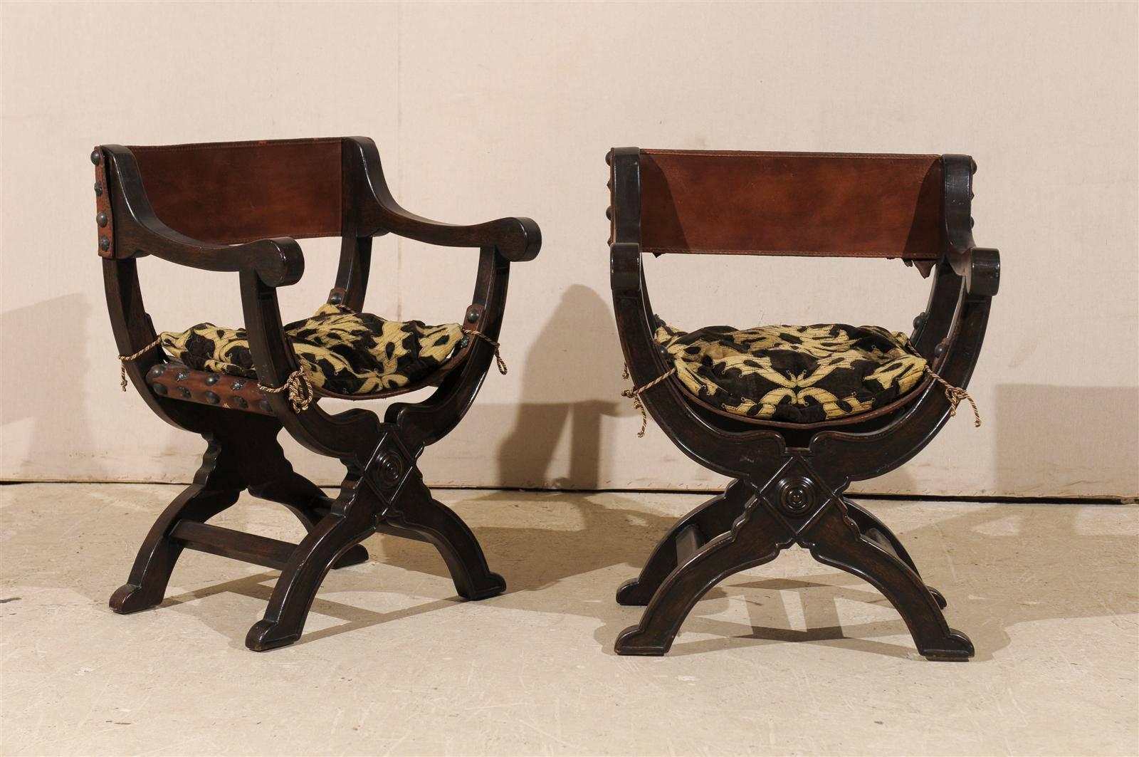 A pair of Italian "Dante," also known as, "Savonarola" style wooden chairs from the 20th century. The seats and backs are done in a nice leather with visible round nail heads. Each chair is adorned with a patterned cushion that