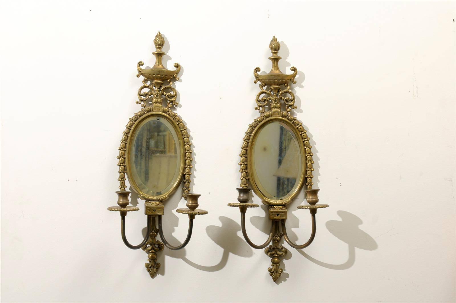 A pair of early 20th century gilded sconces with oval shape and slightly beveled mirror, ornate with two candleholders and topped with a carved fire urn and swag. These sconces are not currently wired.