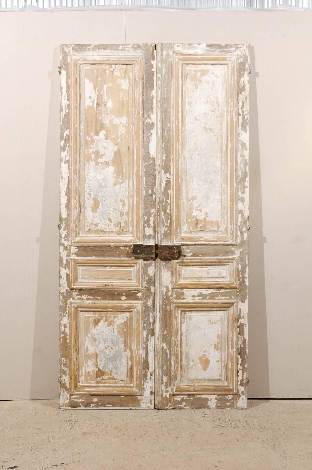 A pair of aged tall French doors from the mid-19th century. These doors have nice old hardware, adorned with acanthus leaf carvings. They have an off white / grey color with some taupe and a lot of wood coming through. These three panel French doors