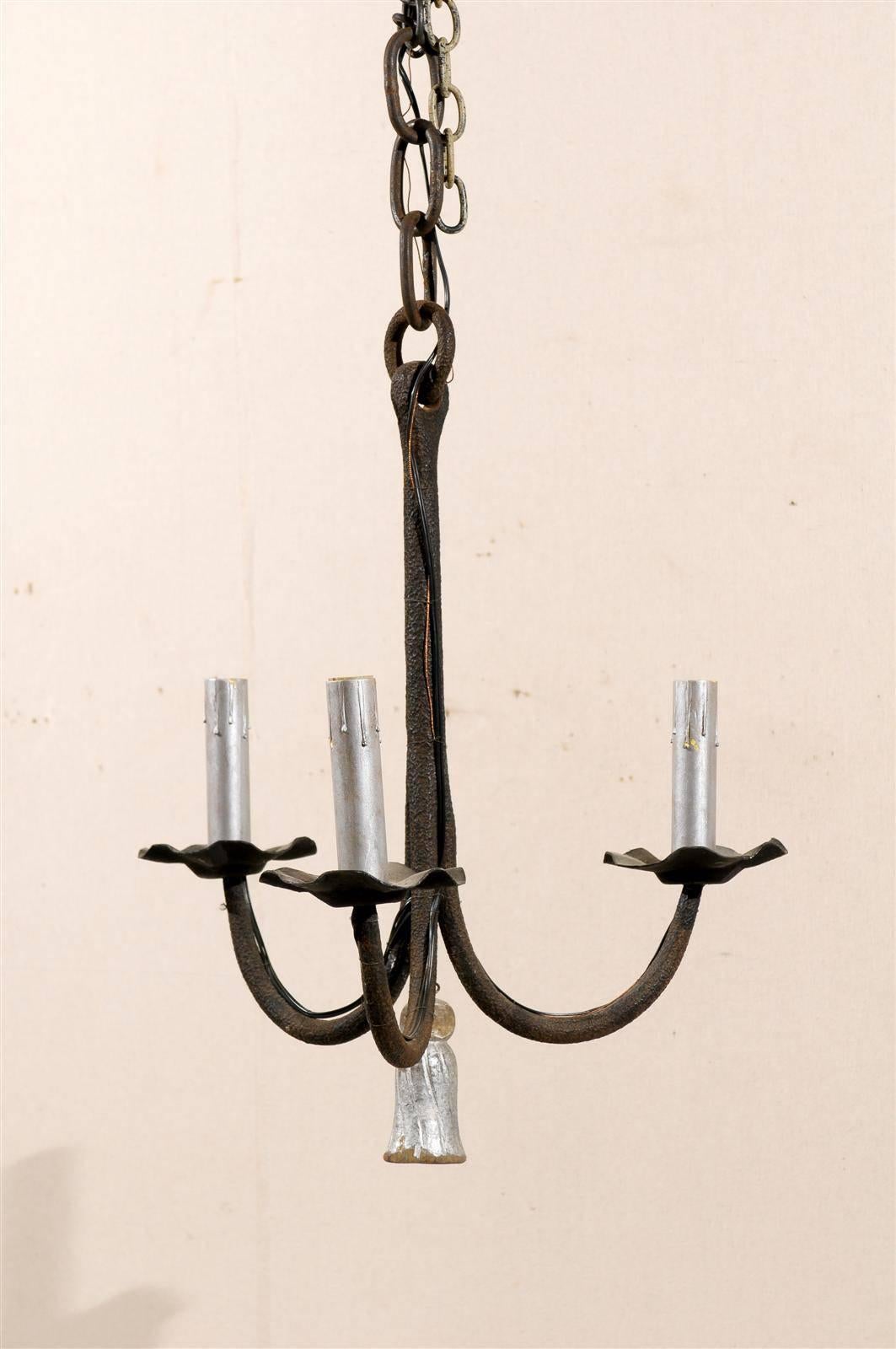 A French three-light iron chandelier. This French chandelier from the early 20th century features swoop arms supporting flower-shaped bobèches and painted candles covers. The lower section of the chandelier is decorated with a silver colored tassel