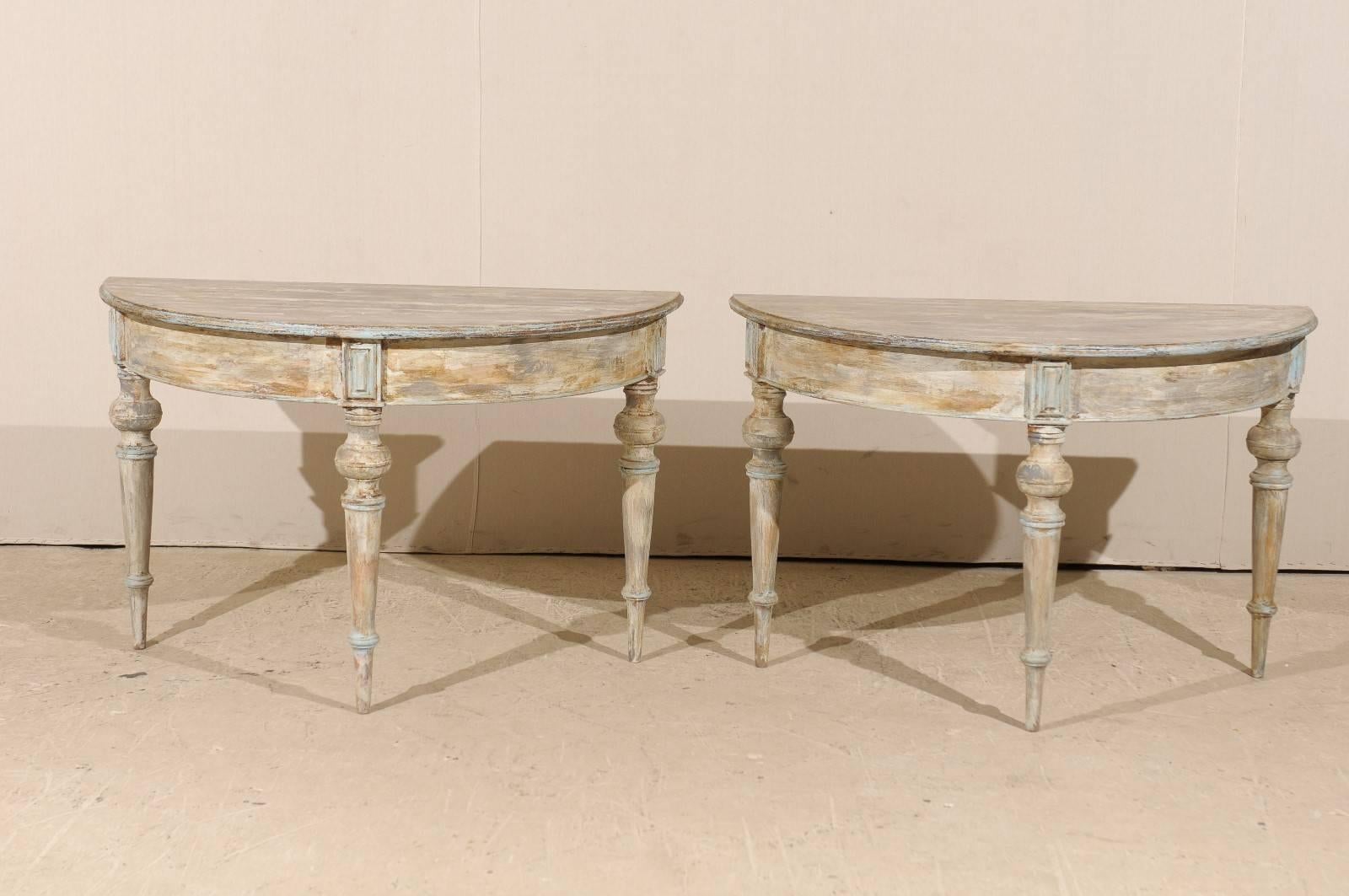 A pair of 19th century Swedish demilune tables. This pair of painted wood demilune tables features half moon tops over circular aprons. The pair is raised on turned and tapered legs. These half moon tables are an overall taupe color with a cream