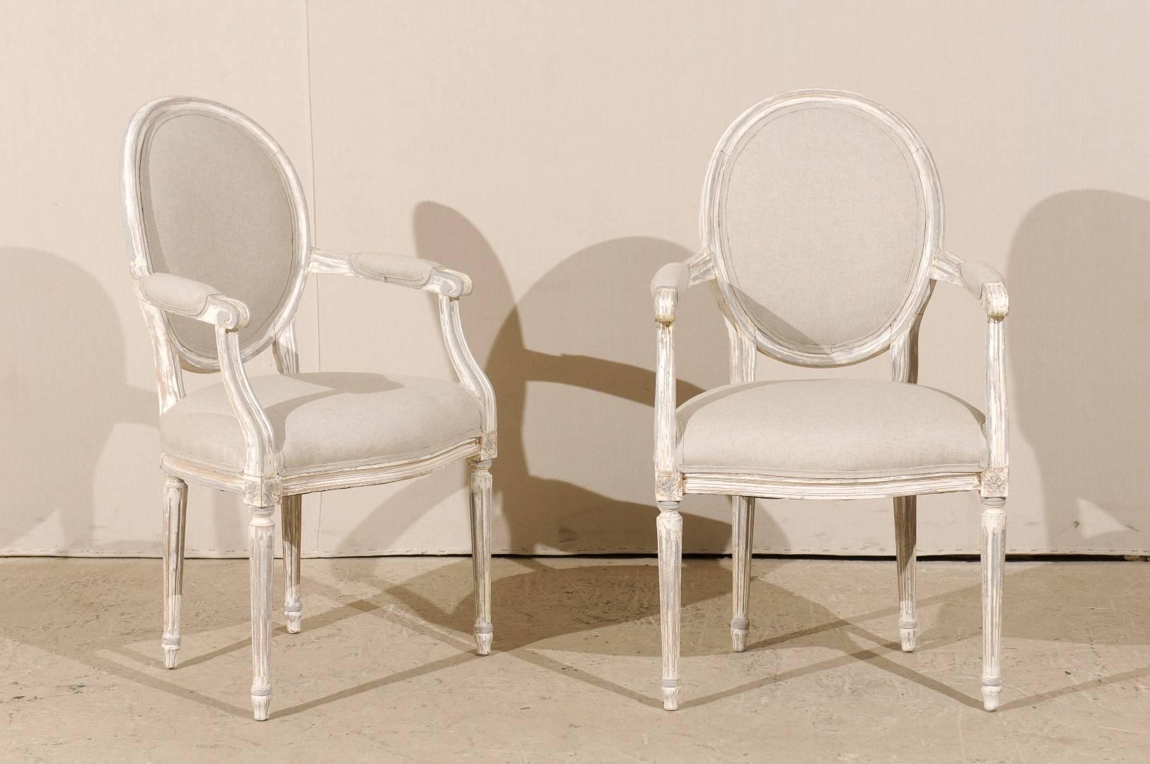 A pair of French Louis XVI style oval back painted wood upholstered accent armchairs. This pair of French chairs features oval backs, scrolled and partially upholstered arms, four fluted and tapered legs as well as rosettes on the knees, typical of