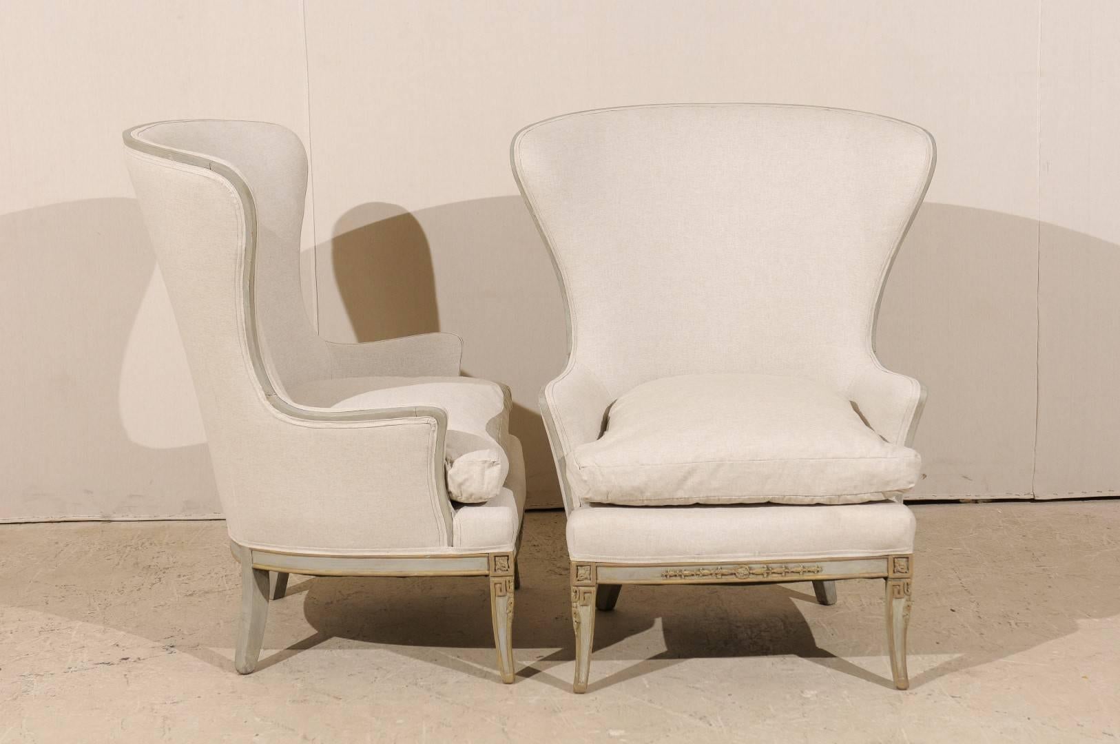 A pair of elegant American vintage carved wood wingback chairs. These painted wood chairs from the mid 20th century have carved details in the center of the skirts and also in the knees. The pair rests on saber legs that have Greek key accents in