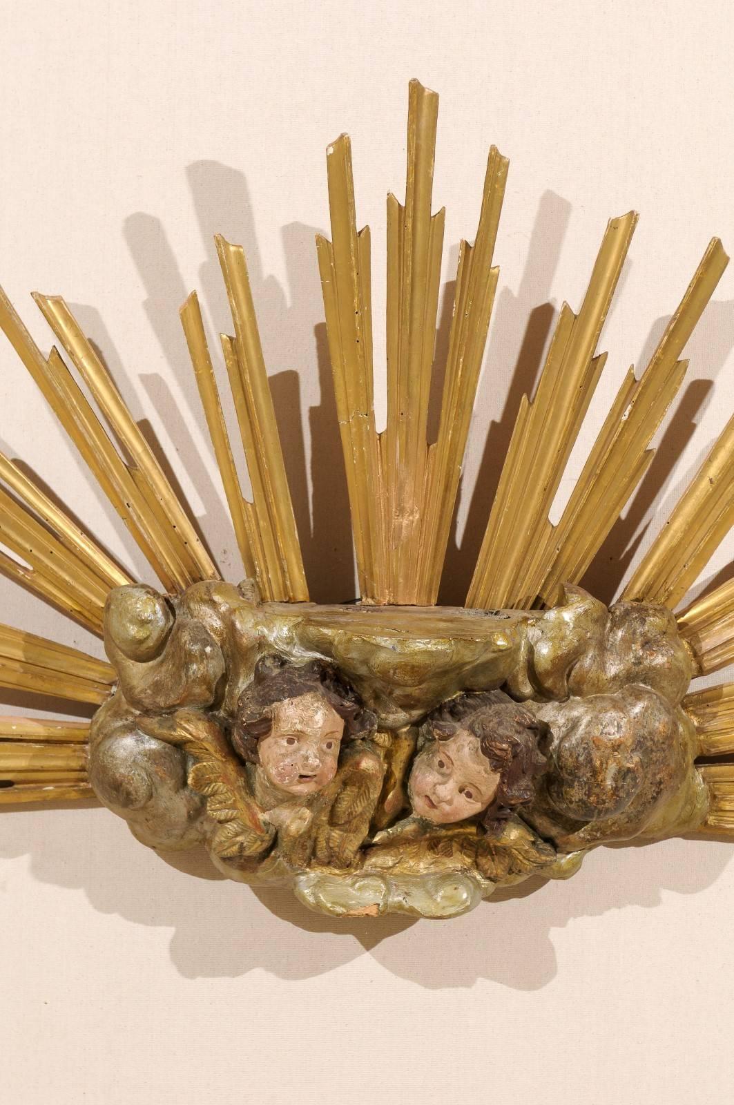 A 19th century Italian giltwood carved gilt sunburst with two carved putti figures in the center. The two putti figures are surrounded by swirling cloud motifs. The gilded rays spring out around the clouds and putti carving creating a half circle