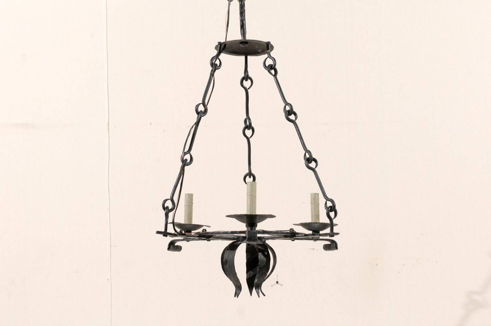A French black forged iron three-light chandelier. This French chandelier from the mid-20th century features metal bobeches and painted candle sleeves at the outer corners. There are four downward projecting leaves decorate the lower section of the