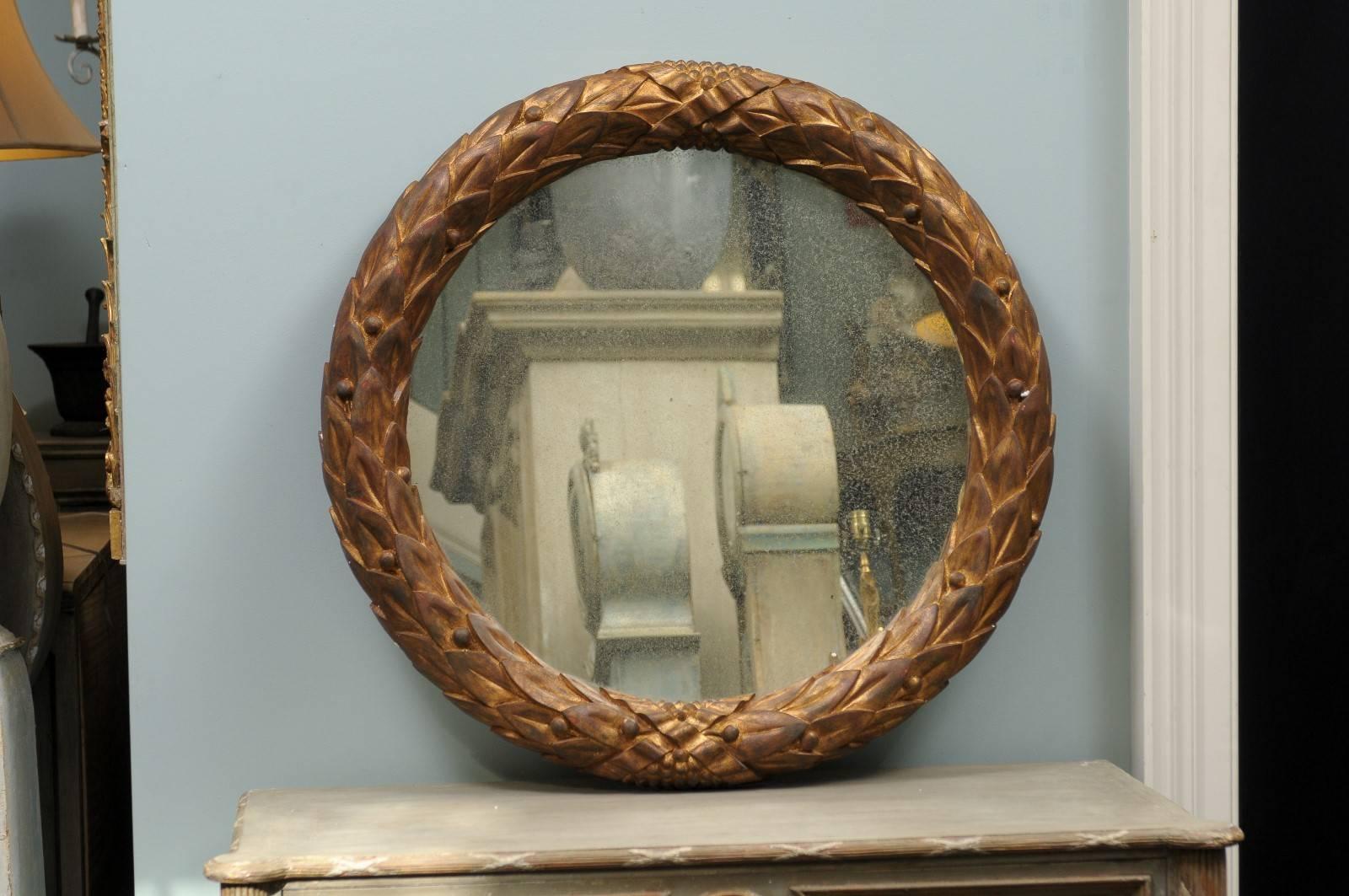 The Garland gilded wood circular mirror. This handmade circular Venetian style mirror features a gilded wood garland motif frame. The central glass has a lightly antiqued finish. The frame shows a gold color with brown accents. This would be a great