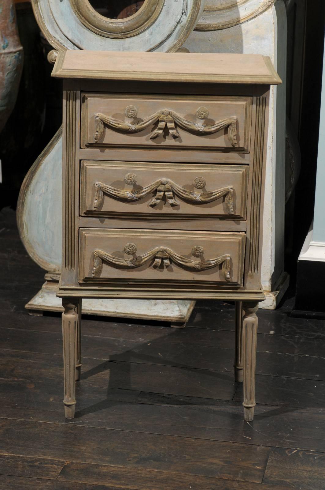 A French small size three-drawer painted wood commode. This little chest from the 1970s features nicely carved swag motifs on each drawer. This piece sits upon slender fluted legs. This three-drawer chest would work really well against a small wall