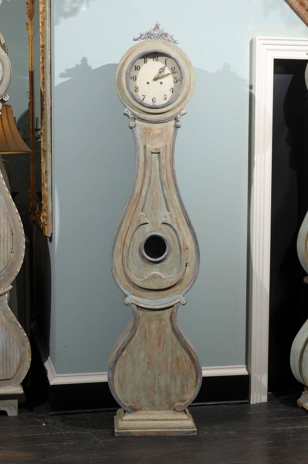 A 19th century Swedish painted wood Fryksdahl clock. This clock features a nice hue of green with aqua, blue and purple accents with some wood coming through. This Fryksdahl's focal point is its carved crest depicting some foliage. The neck and