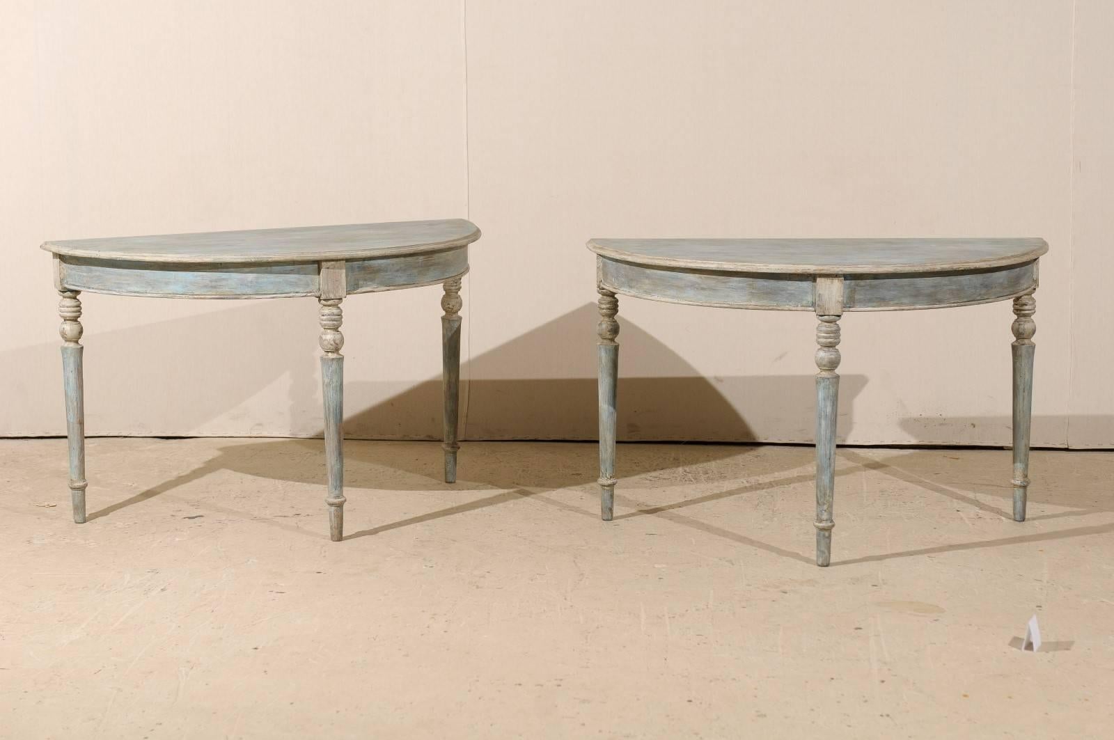 A pair of Swedish 19th century wood demilune tables. This pair of painted wood demilune tables features semi-circular tops over rounded aprons. The tables are raised on three turned and tapered legs. The primary color of these tables is an aqua blue