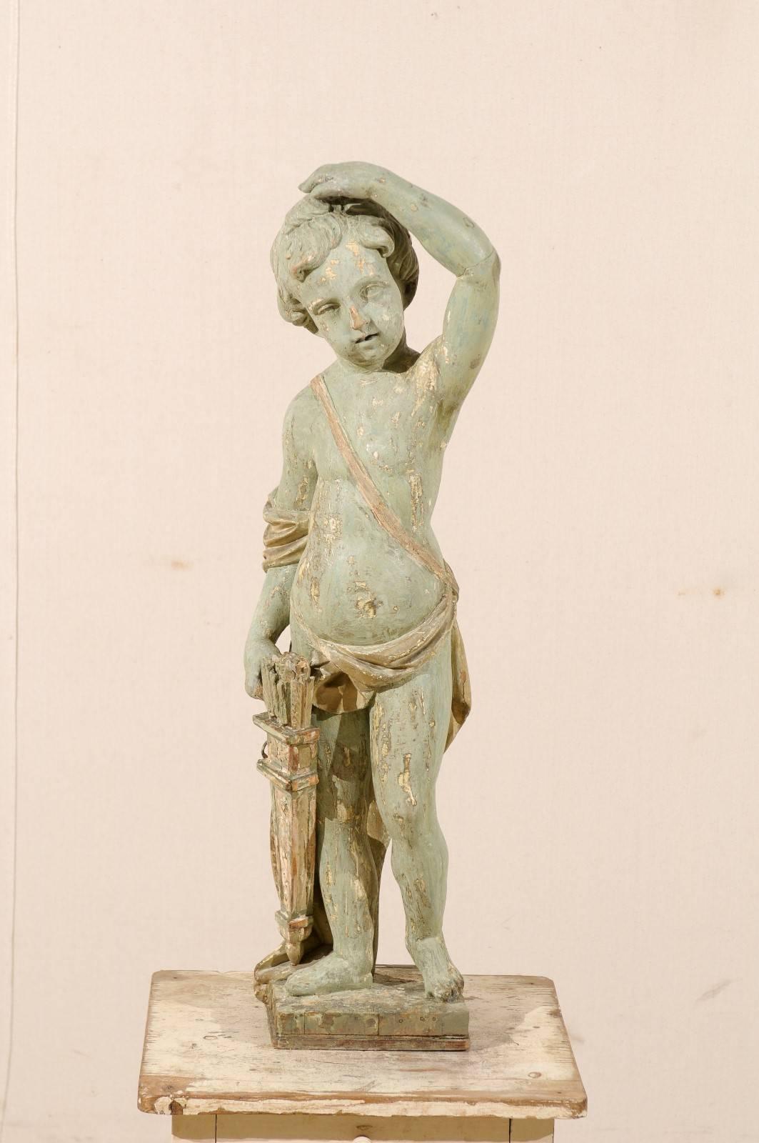 An Italian early 19th century putto figure on wooden stand. This Italian carved and painted wood statue of a putto, commonly known as a Cherub, features a male child raising one hand above his head with the other resting against his quiver with