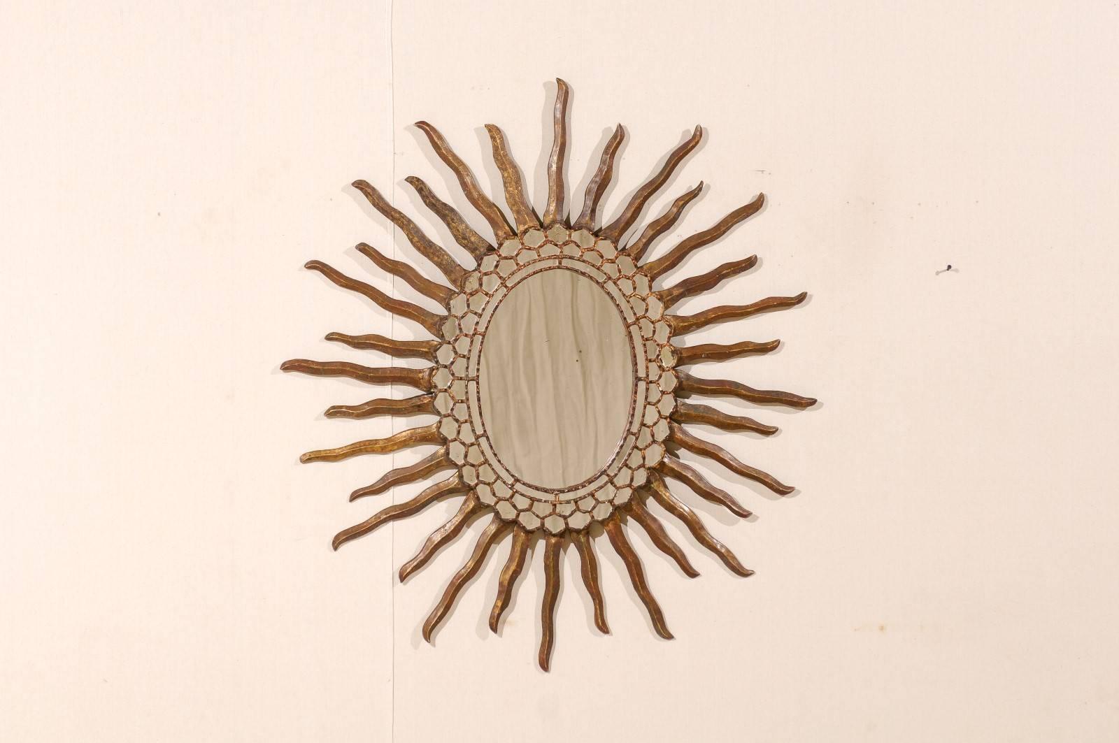 A Spanish antiqued oval shaped sunburst mirror with intricate surround. This Spanish 20th century mirror features a wooden surround in a nice rusty bronze and gold finish. The centre glass is accentuated with a series of concentric rings which are