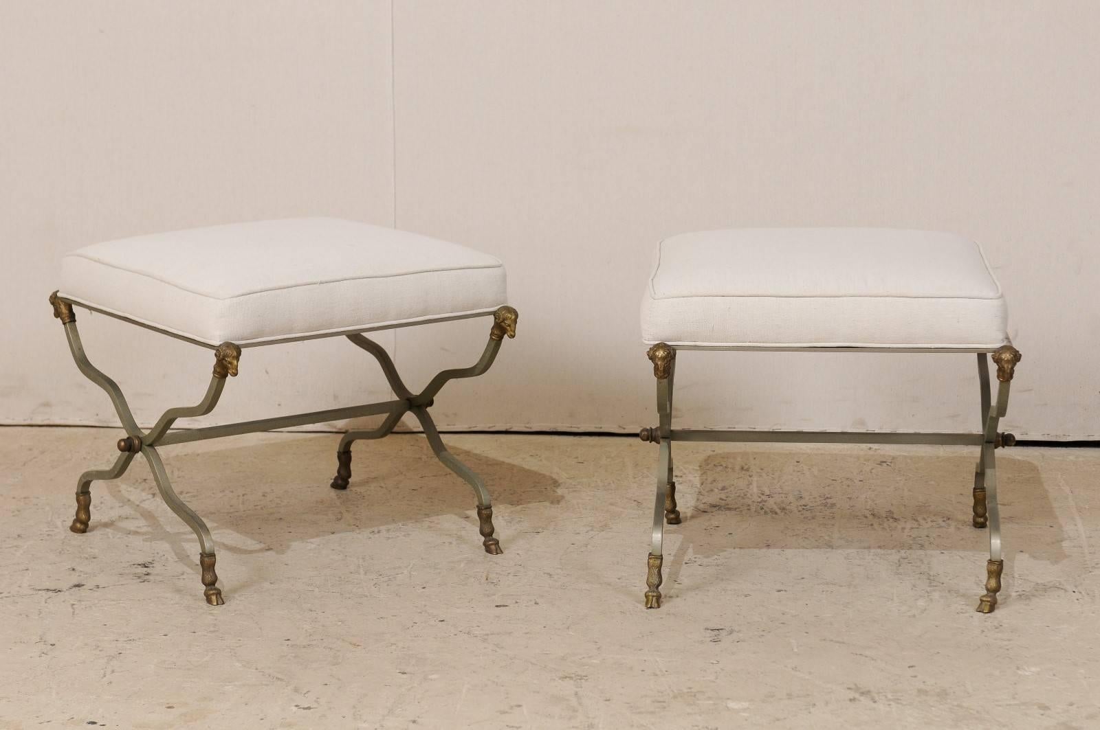A pair of French upholstered x-base stools. This pair of French stools from the mid-20th century feature rams heads at each top leg corner and hoof feet, which are accented with brass. This pair of Mid-Century stools come with a seat cushion which