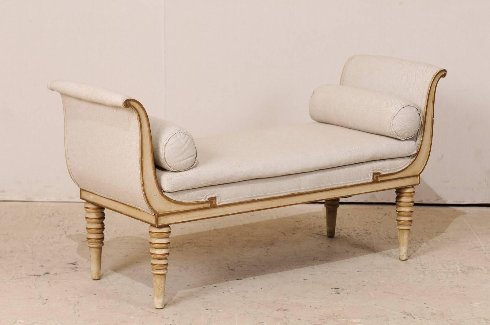 A French Récamier style daybed or bench. This French painted wood bench from the mid-20th century features scrolled arms, turned and tapered legs, and bolster pillows. There is a subtle carved Greek key detail on either side above the skirt. This