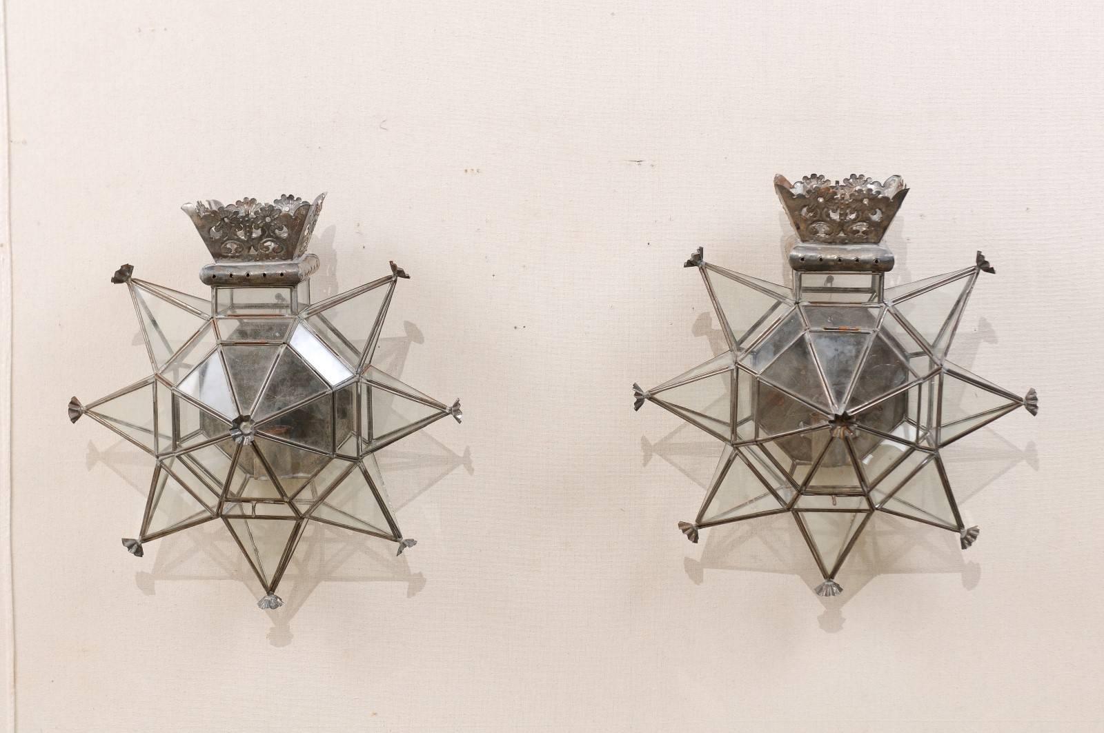 A pair of Mexican silver color star-shaped sconces. This pair of Mexican single candle sconces have been handcrafted from old tin and glass. These Folk Art sconces are both whimsical and noble in style with their glass, star-shaped bodies and floral