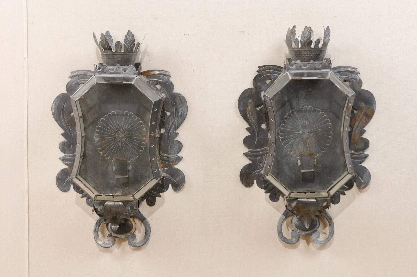 A pair of Mexican Folk Art sconces. This pair of Mexican single candle sconces have been handcrafted from old tin. These sconces feature glass fronts and sides, cut tin accents in a leaf motif, and a leaf and hand-punched crown at top. The interior