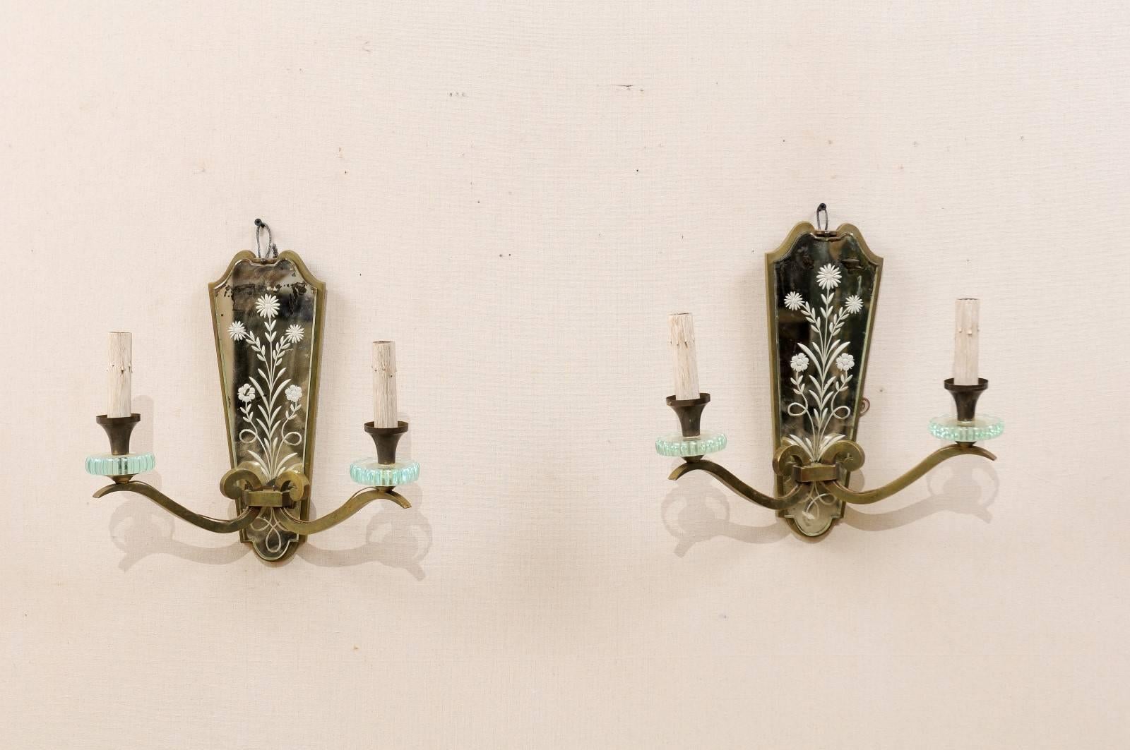 A pair of Mid-Century French two-light Églomisé mirrored sconces. This French pair of sconces, from the mid-20th century, feature a delicately Églomisé or etched mirrored back-plate in a floral motif, with a curved arch top, and straight sides which