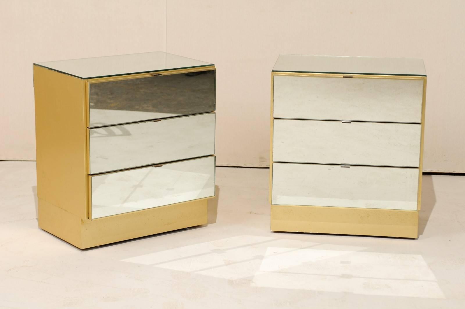 A pair of modern style mirrored chests. This vintage pair of American three-drawer chests feature clean, modern lines with mirrored panels across each of the three drawers and tops. The body is gold in color and made from melamine or similar type