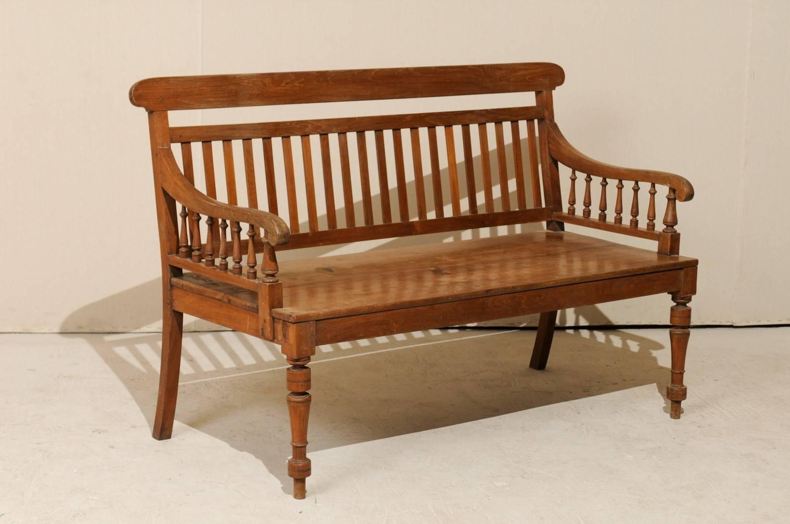 A British Colonial teak wood bench. This British Colonial style teak wood bench is from the mid-20th century, India. This sofa bench features turned front legs, a vertical slat back, bannister style arms and curled knuckles. Teak is a tropical