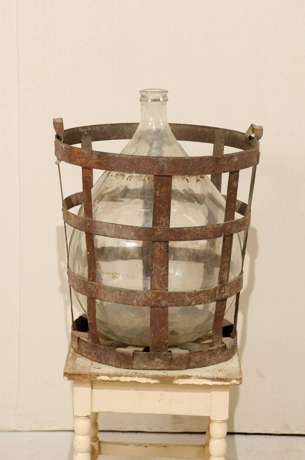 A Mid-Century French vintner iron basket with demijohn wine bottle. This is a French vintner iron-strap, two handle basket containing a large sized, clear, demijohn wine bottle. The basket is nicely aged. This French vintner basket and demijohn
