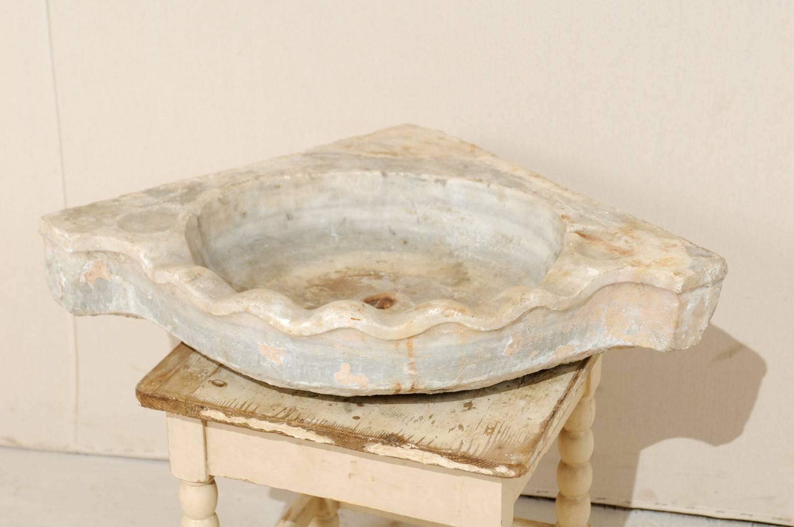This is a European 19th century marble corner sink. This 19th century marble sink is designed to fit into a corner with a curved front. The coloring is primarily neutral tones of porcelain and alabaster. It is carved beautifully, with a scalloped