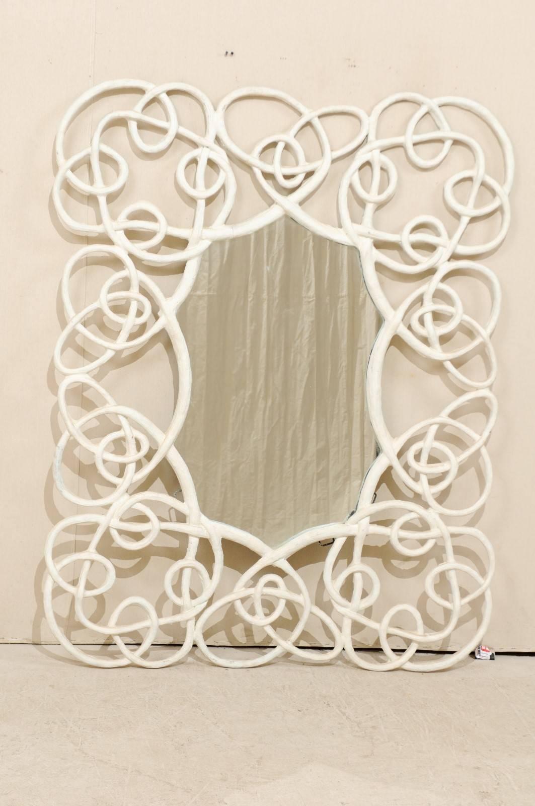 A large modern mirror. This mirror features a twisted, vine like surround which has been painted an off-white color. It is made of a composite material with a custom finish and has an overall rectangular shape. This whimsical mirror would be great