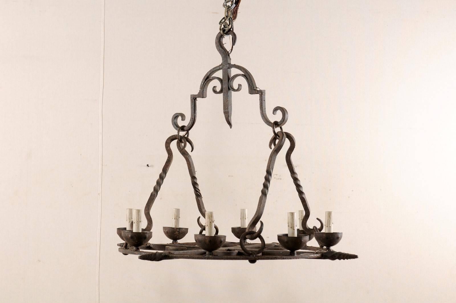 A French Mid-Century eight-light chandelier. This French chandelier from the mid-20th century of forged iron has a flattened, circular shaped body which supports the eight iron bobèches and candle covers around its perimeter. The lower section is