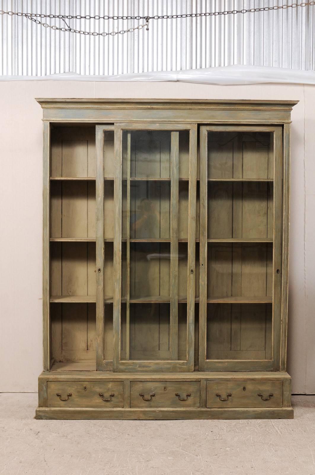 A French late 19th century large painted wood bookcase with sliding doors. This French cabinet, circa 1880, features three sliding glass doors with adjustable wood shelving behind, over three outset bottom drawers. It has a molded cornice with rope