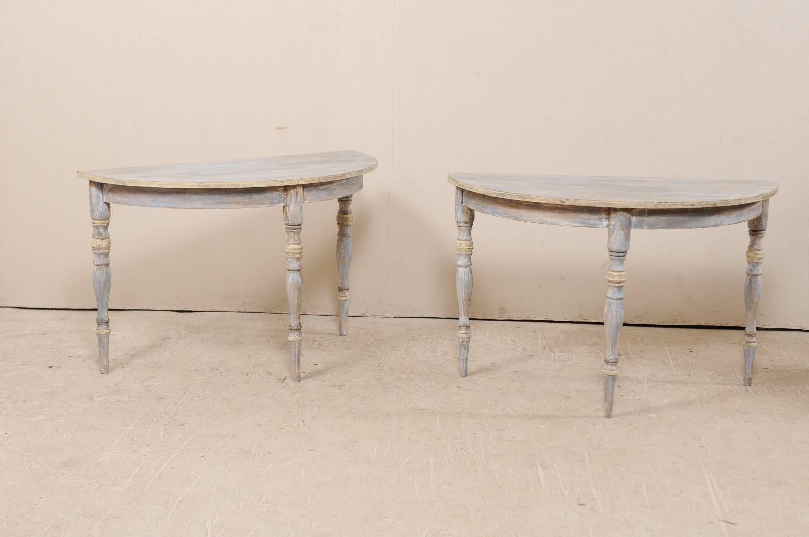 A pair of 19th century Swedish demilune tables. This pair of painted wood demilune tables features half moon tops over round aprons. These demilune console tables are raised on three turned and slightly tapered legs. They are primarily blue in color