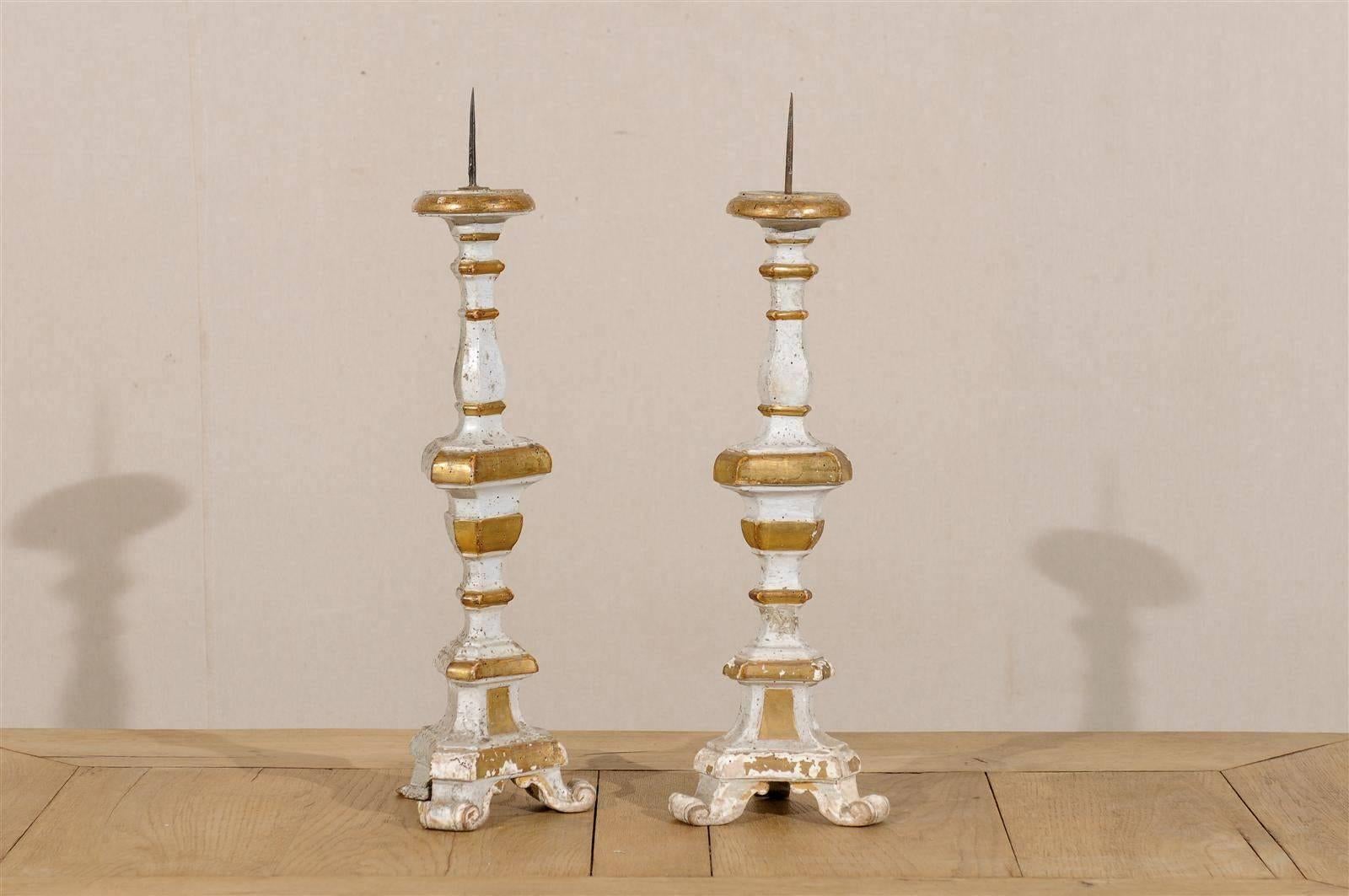 A pair of 19th century Italian candlesticks. These wooden candlesticks are painted a soft cream and white color. Each candlestick is accented with gold gilding. These candlesticks rest on tripod feet. This pair of Italian candlesticks would look