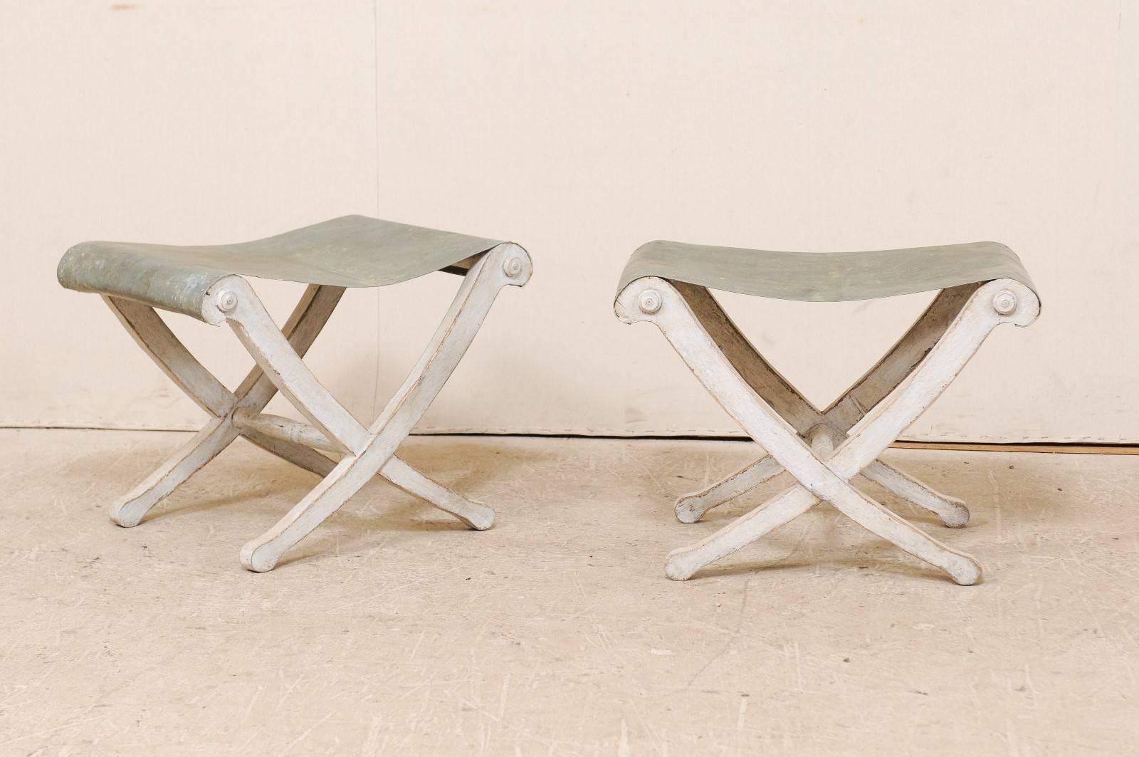 A pair of Italian carved wood stools with zinc seats. These vintage Italian stools feature whimsical zinc seats and carved wood bases with lightly swagging x-crossed legs at either side. The legs are simple with gently rolled feet and tops. The zinc