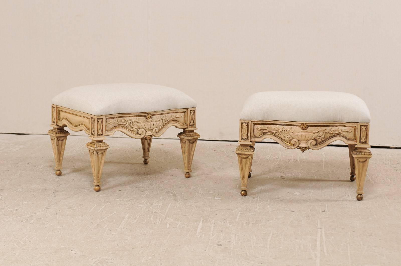 A pair of vintage carved wood and upholstered stools. This pair of American stools are richly carved ashwood in an Italian style. The skirt is valanced with a stylized urn and scroll motif at their centers. The stools are raised upon exquisitely