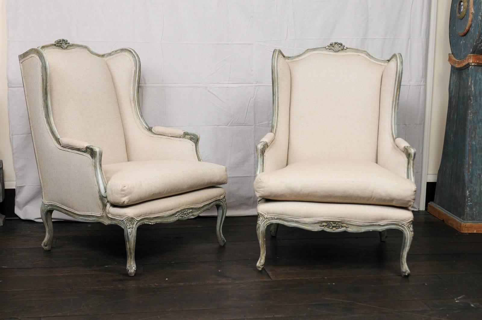 A pair of mid-20th century French painted wood and upholstered wingback chairs. This pair of French midcentury winged back chairs have nice wide seats and manchette arms. The chair's wood carvings feature scrolled knuckles, scalloped skirts,