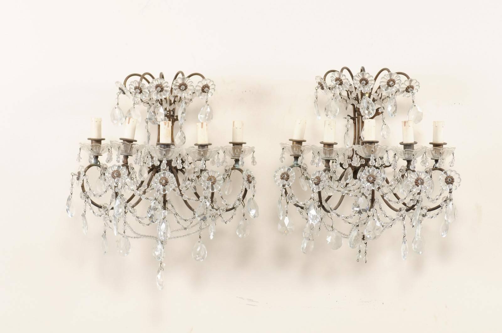 A pair of Italian crystal five-light sconces from the mid-20th century. This pair of Italian crystal sconces each feature five lights set within scrolling metal armature, adorn with delicately swagged crystals, beading and crystal flowers