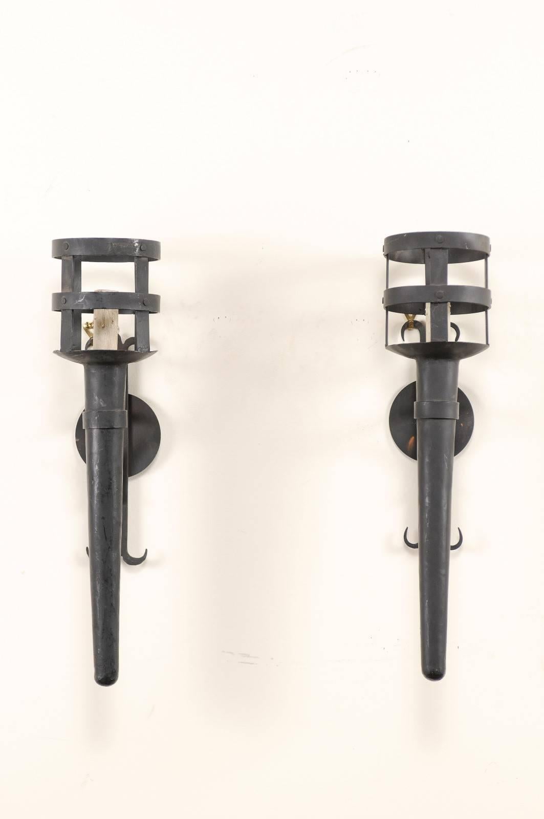 A vintage pair of American iron torch sconces. This pair of American single-light sconces have long, vertical torch-style arms which are supported by their round backplates with fleur-de-lis stylized bars fixed vertically through their centres. The