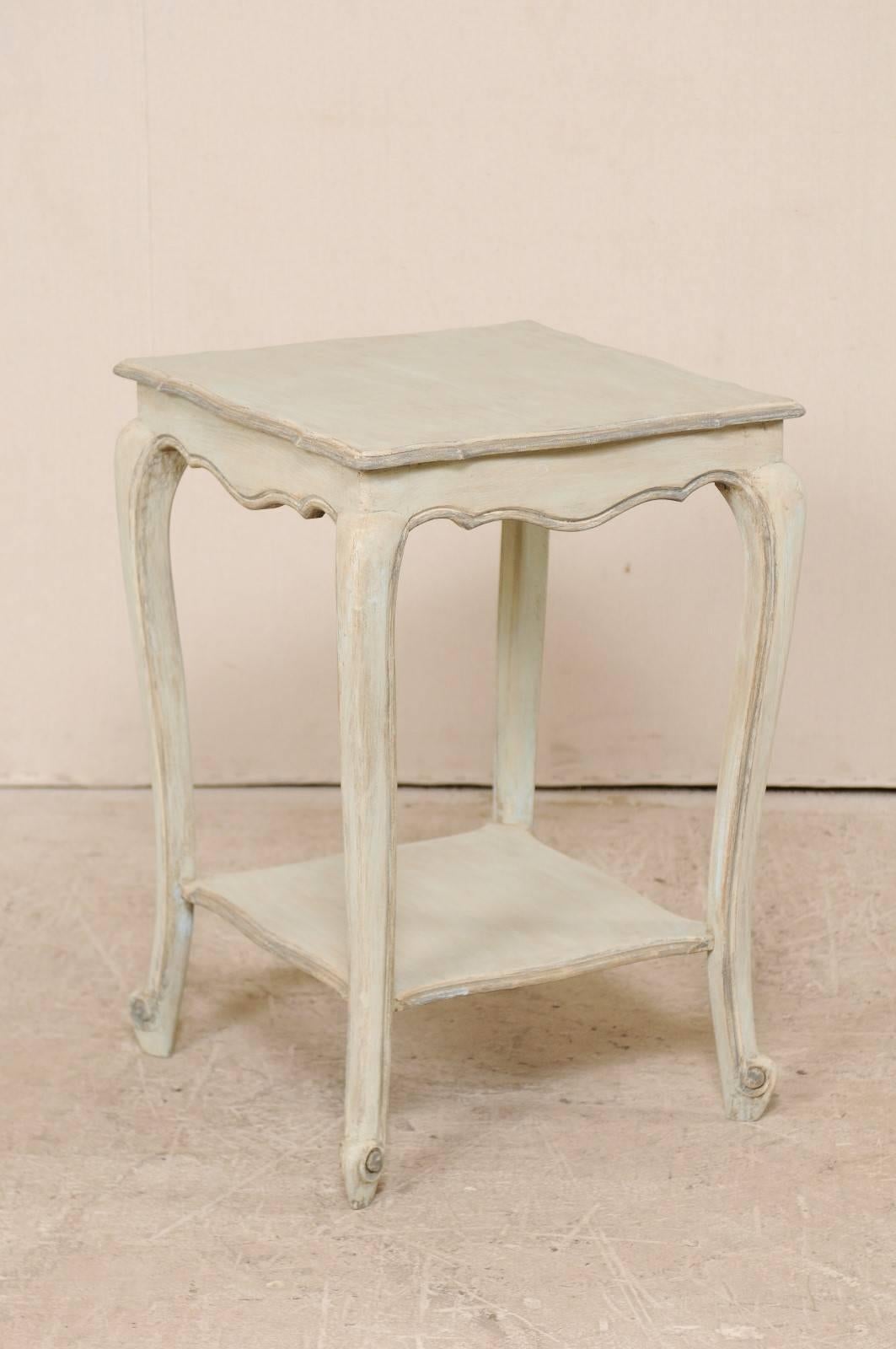 A French, early 20th century painted wood side table. This antique French end table features a square-shaped top with scalloped edges over a scalloped skirt. There is a lower shelf and four slender cabriole legs. This sweet little side table is a