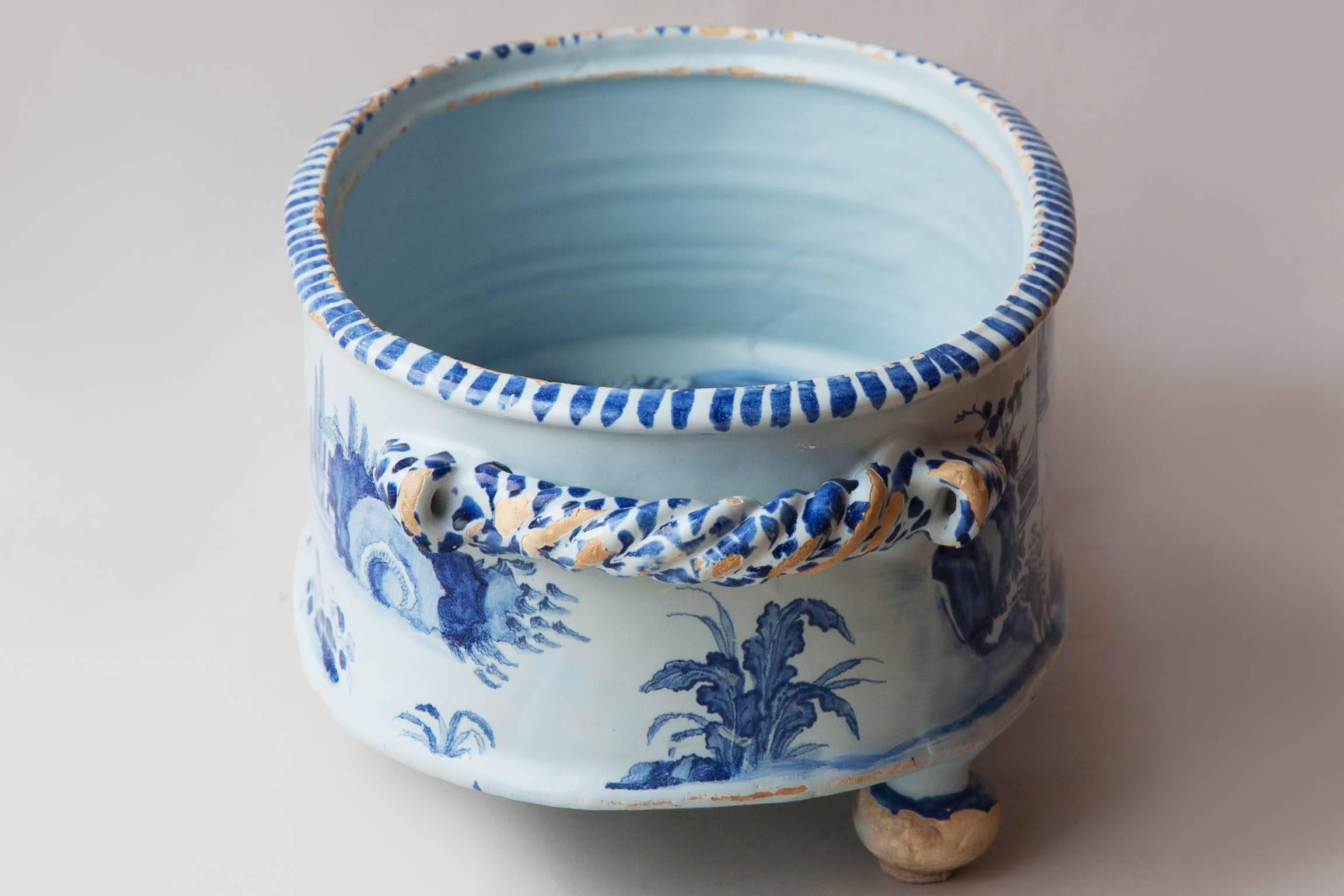 The oval body standing on three round feet, with twisted rope handles. Depicting Chinamen in a garden setting  with plants and trees painted in blue on a pale bule glazed white background late 17th Century, Circa 1680.

Chips and scratches to the