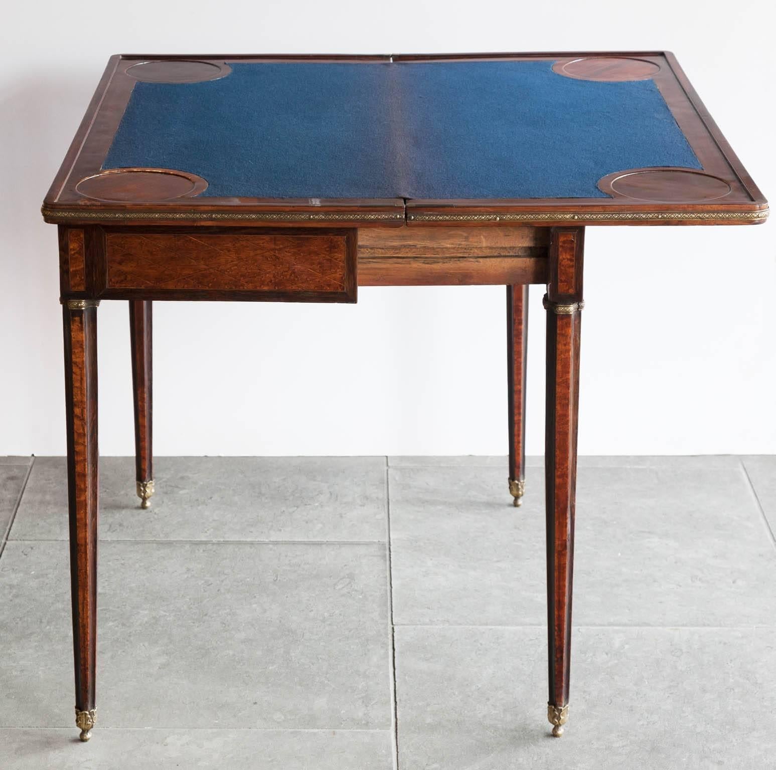 Having a geometrical marquetry hinged top with enclosing a blue baize game surface. Cast brass edging around the folded top.
Standing on slender fluted legs with brass rings, terminating in brass caps cast in the shape of oak leaves. The Table is