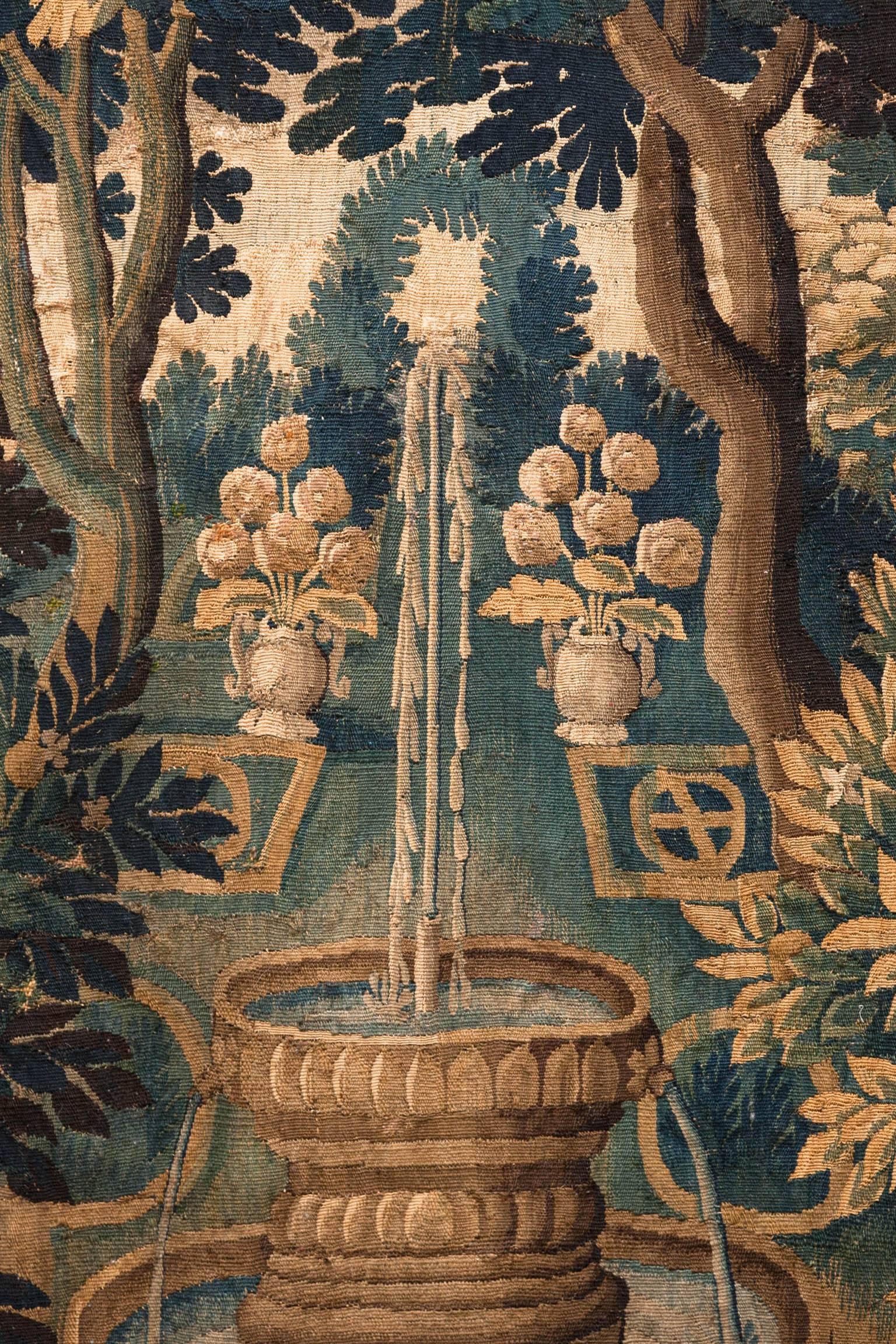 Verdure tapestry with two birds perched on plinths either side of two fruit trees which surround a fountain in an 18th century garden in the foreground.
In the background two urns hold flowers. With a third bird perched on top of the branch of a