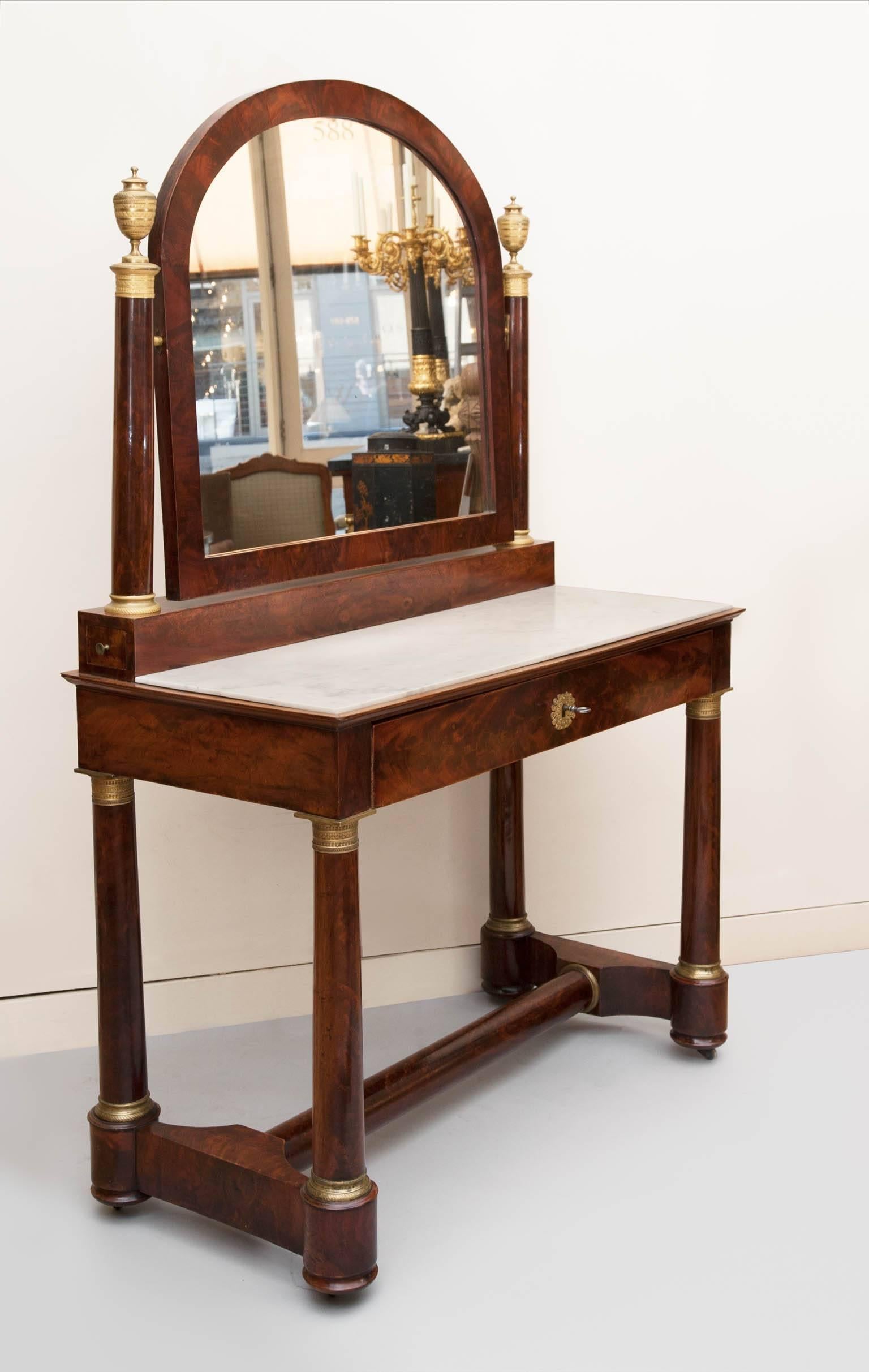 With a hinged mirror framed in mahogany, supported by two columns surmounted with fine quality gilt bronze urns. The mirror rests on a mahogany plinth with two sideways opening drawers, this on top of a table on four flame mahogany column legs, all