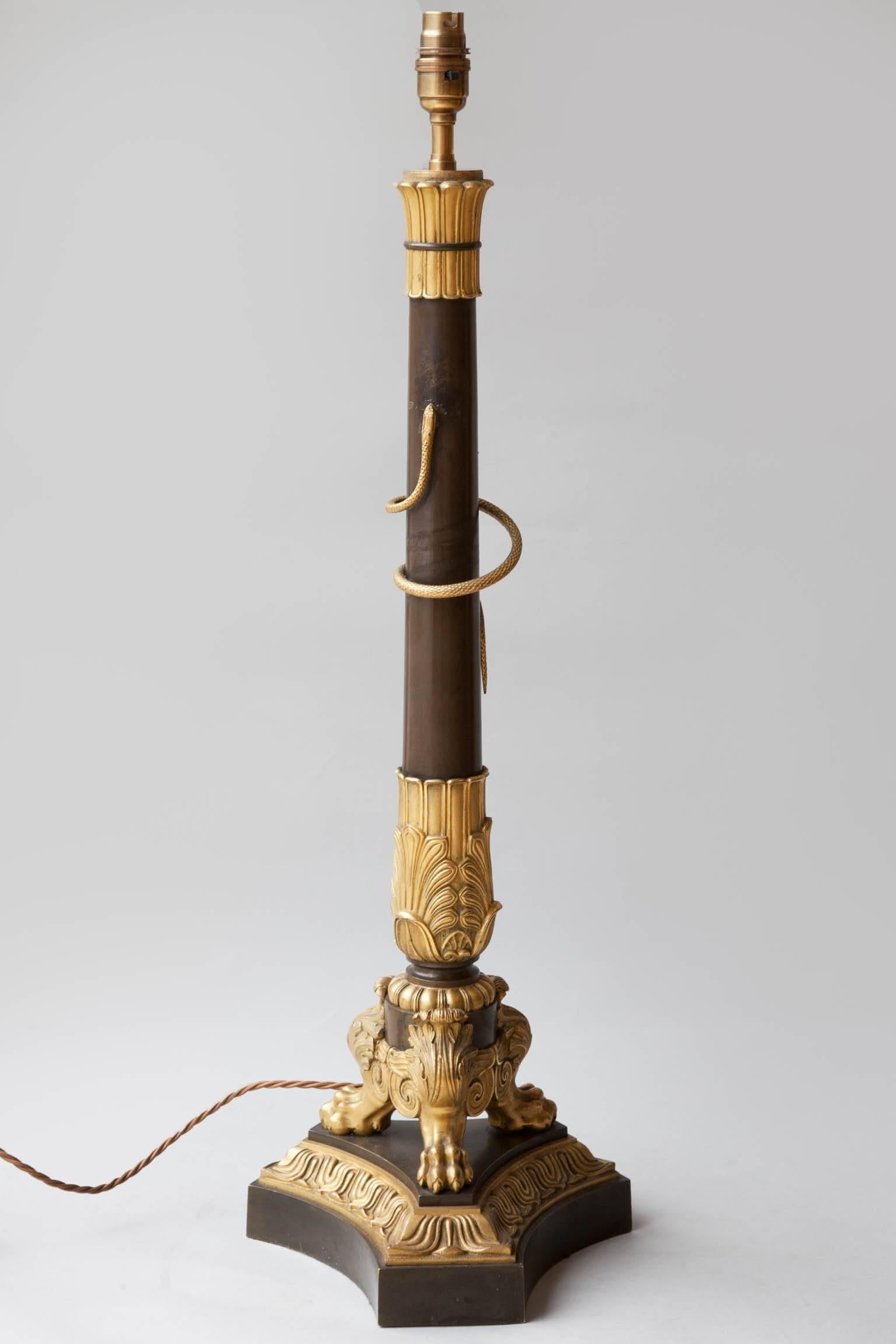 Early 19th century candlestick converted to a table lamp. Cast gilt leaves surround the top and bottom of the patinated column, supported on three gilt bronze paw feet mounted on a concave tripod base. A gilt bronze snake is attached to the central