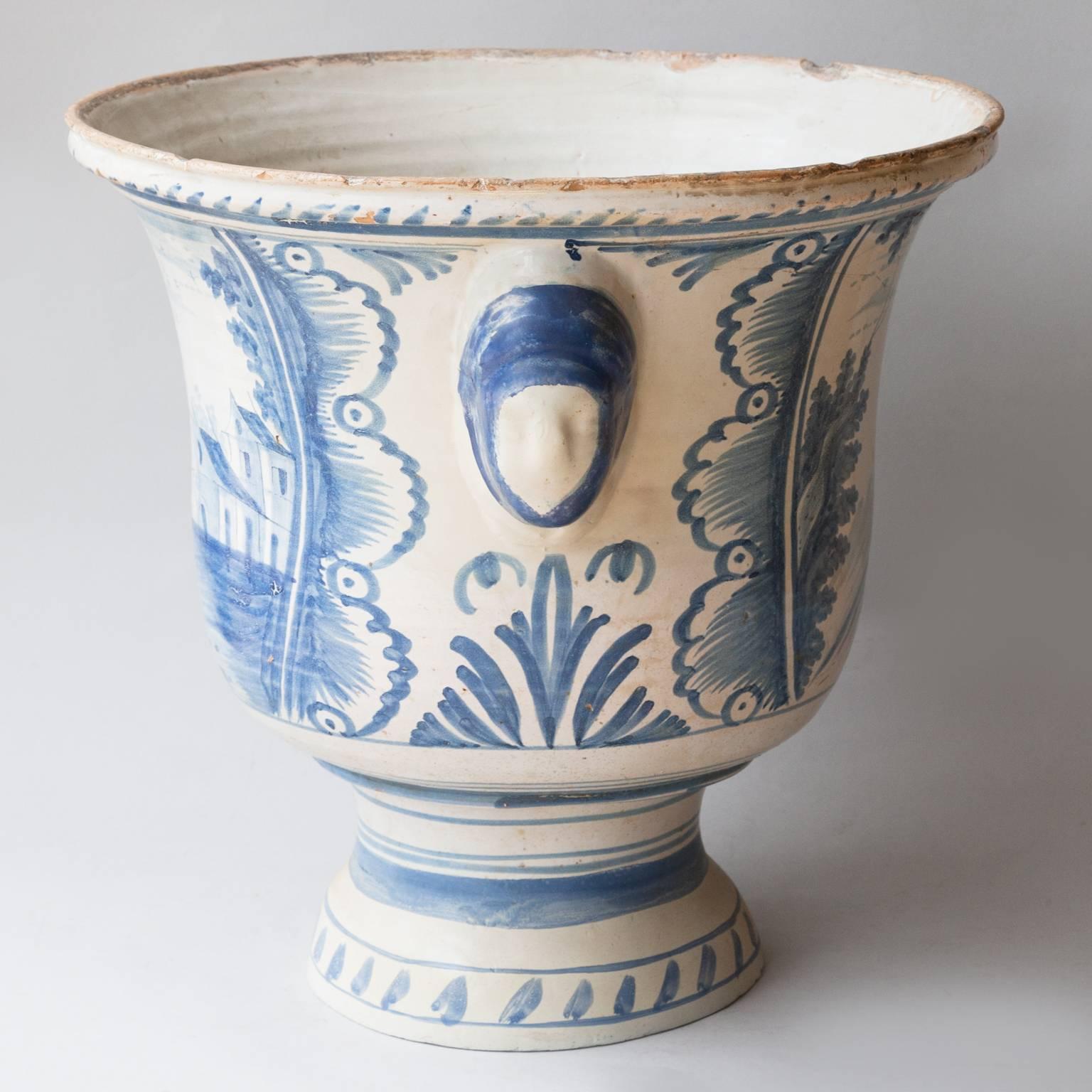 Blue and white glazed faïence large jardiniere decorated with pastoral scenes, raised on a plinth base with
handles in the shape of masks. 
Restoration to the base and handles. Rouen, France, circa 1800.
 