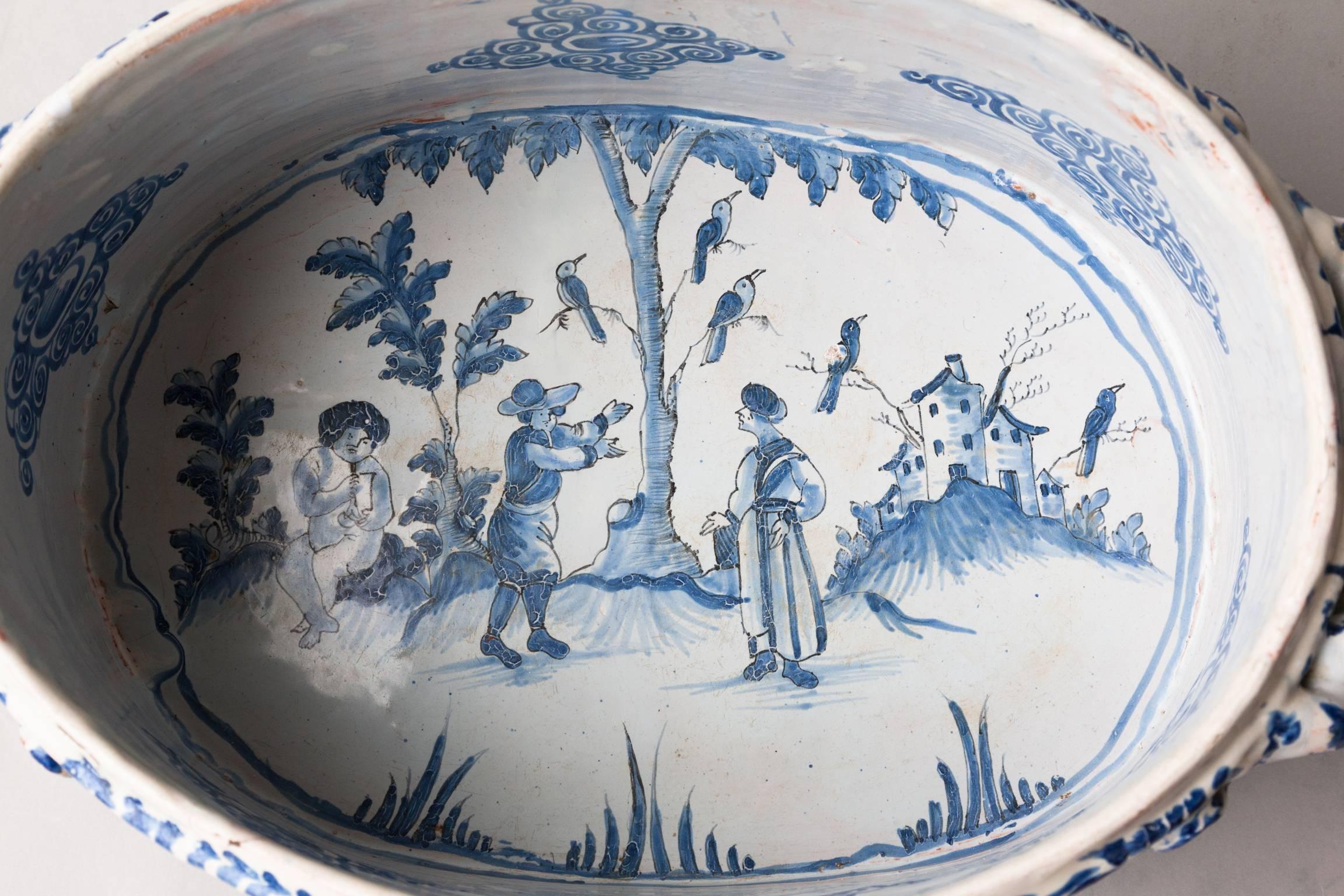 The oval body standing on three round feet, with twisted rope handles. Depicting oriental figures and a musician in a landscape setting 
With a building, plants and birds in a tree, painted in blue on a pale bule glazed white background late 17th